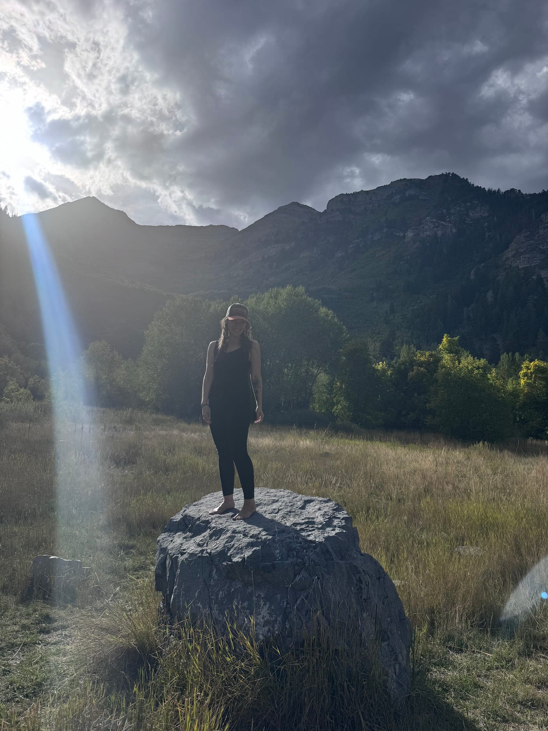 Rachel standing barefoot on a boulder in a mountain meadow, a beam of light falling beside her